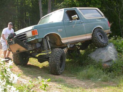 1990 Ford Bronco - Green Ford Bronco with white top and tan lower stripe flexing with its rear passenger tire up on a large rock at the edge of a grassy field…