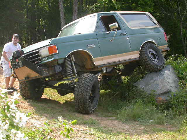 Green Ford Bronco flexing with rear tire on a boulder beside a grassy treeline