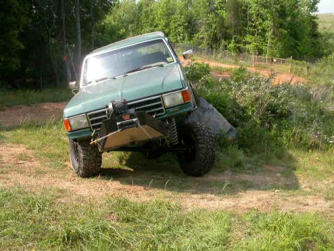 1990 Ford Bronco - Front three-quarter view of a lifted green Ford SUV flexing off-camber on a grassy trail bank, its front axle articulated with one mud tire…