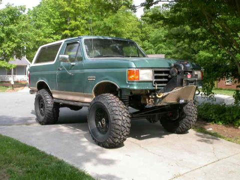 1990 Ford Bronco - Green Ford Bronco with a white fiberglass removable top and tan lower body stripe, parked on a residential driveway in a three-quarter fron…