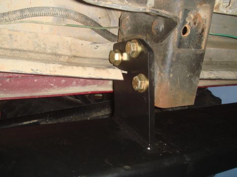 1992 Ford Bronco - Close-up under a truck's rear quarter showing a black steel bumper mounting bracket bolted to the frame rail with three yellow-zinc flange…