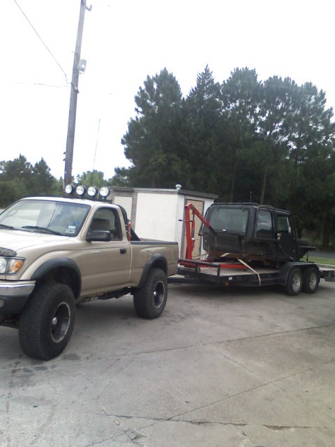 1982 Ford Bronco - A Toyota Tacoma tows a trailer carrying a disassembled vehicle body, with a utility building in the background.