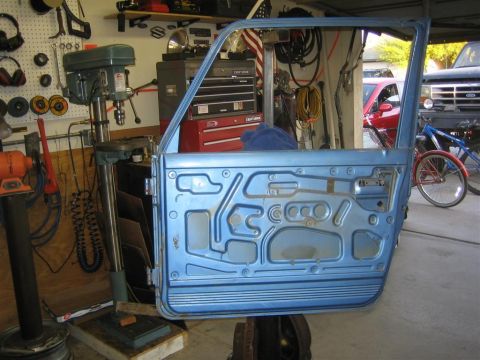 1987 Suzuki Samurai - A blue car door is shown with its interior bracing visible, in a workshop setting with tools and other vehicles in the background.