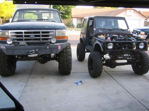 1987 Suzuki Samurai - Two modified off-road vehicles, a black Suzuki Samurai and a dark blue Ford truck, are parked side-by-side in a driveway.