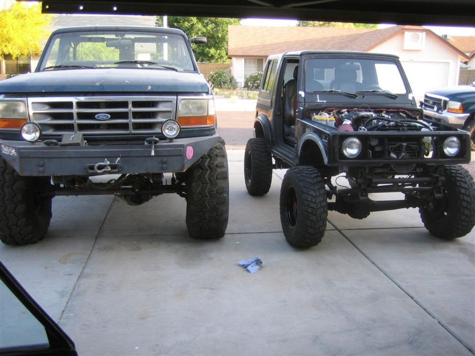 Black Suzuki Samurai with hood open, front view, parked next to a Ford truck
