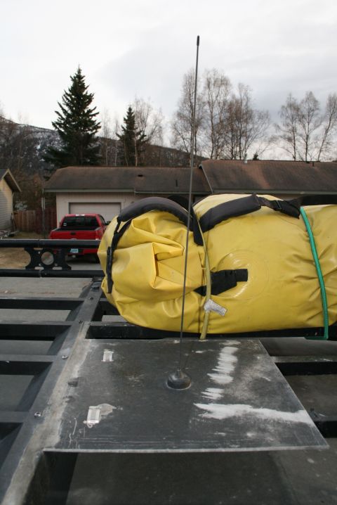2003 Jeep TJ - A yellow inflatable raft is strapped to a trailer, with a red pickup truck parked in front of a garage.