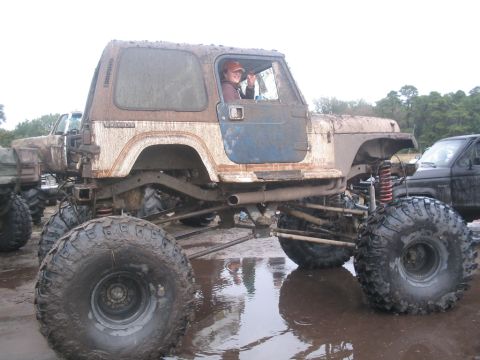1990 Ford Bronco - A heavily modified Jeep CJ-7 navigates a muddy terrain, with another vehicle partially visible to the right.