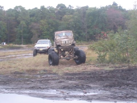 1990 Ford Bronco - A heavily modified off-road vehicle with oversized tires sits in a muddy area, with another white SUV visible in the background.