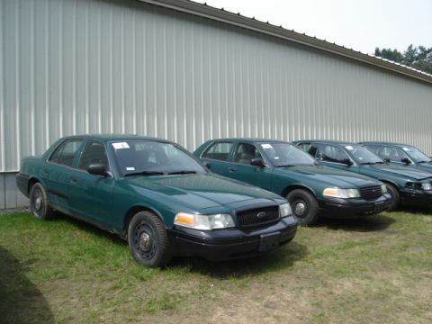 2003 Ford Crown Victoria - Several green 2003 Ford Crown Victoria sedans are lined up in front of a metal building.