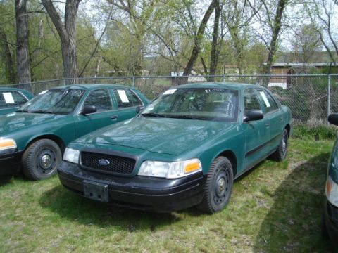2003 Ford Crown Victoria - Several green Ford Crown Victorias are parked in a grassy lot under a cloudy sky.