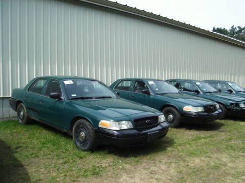 2003 Ford Crown Victoria - A line of dark green Ford Crown Victoria sedans are parked on a grassy area in front of a corrugated metal building. The front-most vehicle…