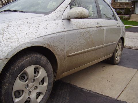 1996 Ford F-150 - A silver sedan is heavily covered in mud, with the front driver's side tire and fender prominently displayed. The vehicle is parked on a co…