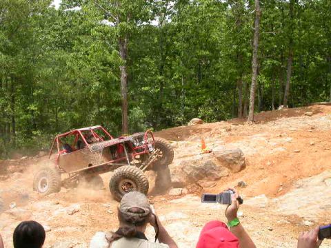 1990 Ford Bronco - A custom red off-road buggy with large tires navigates a rocky, dirt incline in a wooded area. Spectators with cameras are visible in the f…