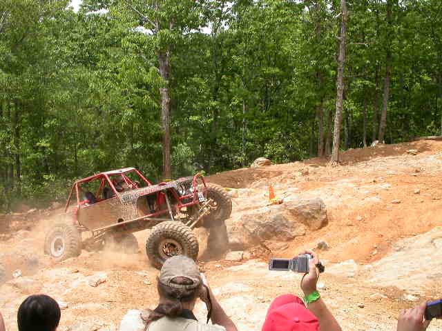 Red custom off-road buggy climbing a rocky hill in a forest with spectators filming.