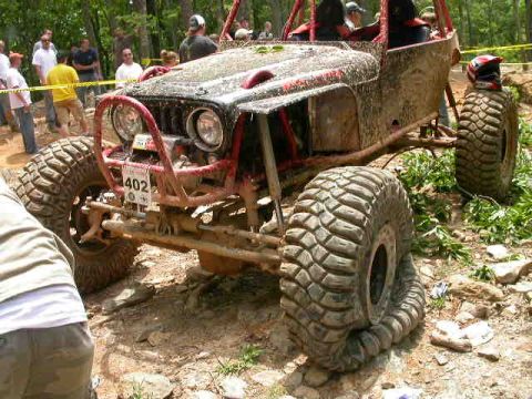 1990 Ford Bronco - A heavily modified, mud-covered Jeep Wrangler is parked on a dirt path with large, aggressive tires. The vehicle has a red roll cage and a…