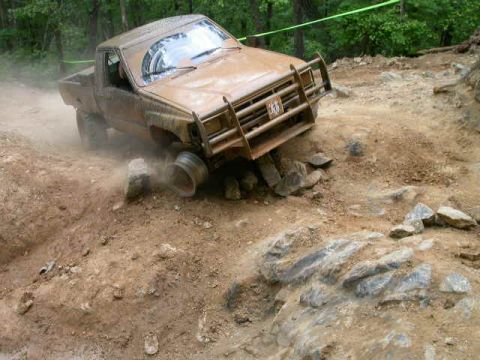 1990 Ford Bronco - A brown, mud-covered pickup truck is navigating a rocky, off-road course, with its front passenger-side wheel lifted off the ground.