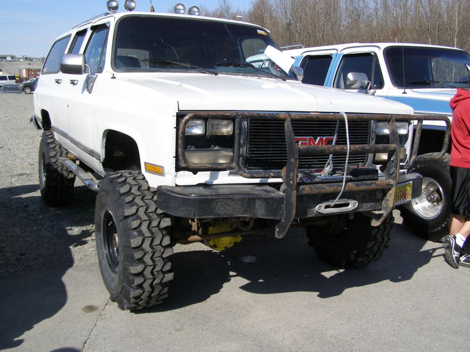 Lifted white GMC SUV with off-road tires and brush guard, parked on pavement.