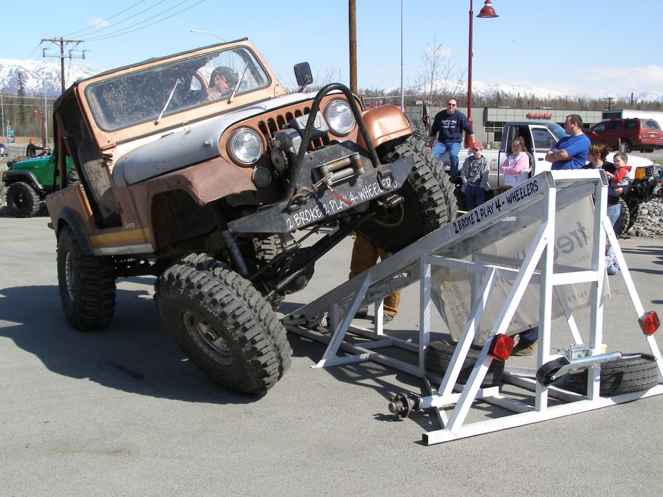 Rusty orange and silver Jeep CJ angled on a metal ramp with people watching.
