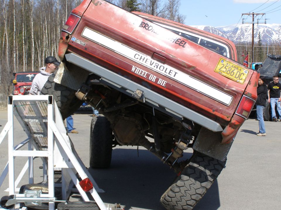 Heavily modified red Chevrolet pickup truck tilted on a trailer, showing large off-road tires and suspension. Tailgate reads "CHEVROLET WHEEL OR DIE".