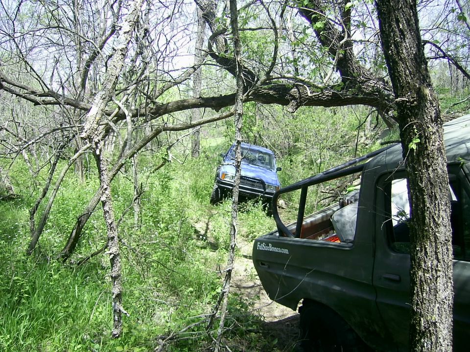 Two off-road vehicles navigating a wooded trail, one blue SUV in the background and a dark-colored truck in the foreground