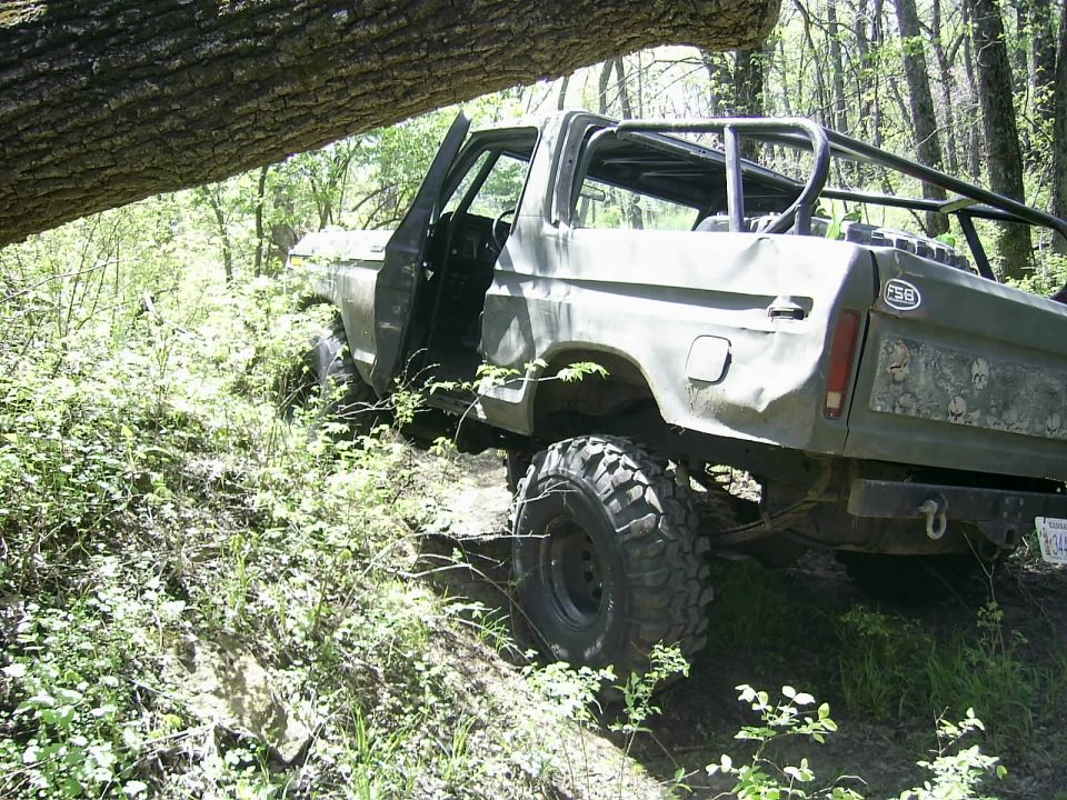 Olive green 1978 Ford Bronco with roll cage, off-road tires, and open doors, tilted on a wooded incline