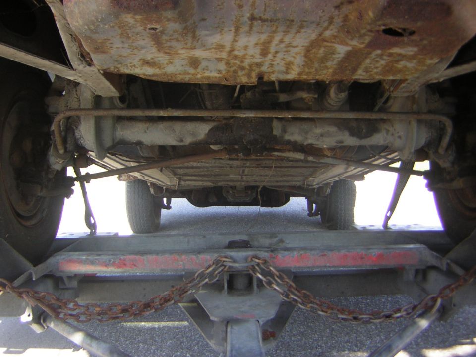 Underside view of a vehicle being transported on a trailer, showing rusty undercarriage and suspension components