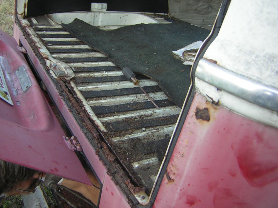 Interior of a vintage vehicle cargo area with a dark mat over ribbed flooring, red exterior, and rust.