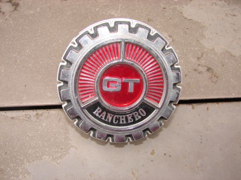 1973 Ford Ranchero - A chrome gear-shaped 'GT RANCHERO' emblem with a red radial-sunburst background and 'GT' in the center, resting on a dusty concrete surface.