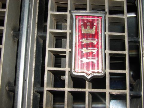 1973 Ford Ranchero - Close-up of a red shield-shaped grille emblem showing a crown above three walking lions, mounted in the center of a dusty egg-crate grille.…