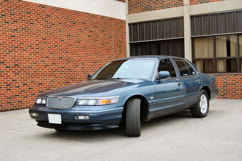 1985 Mercury Grand Marquis - A blue sedan is parked in front of a brick building with large windows.