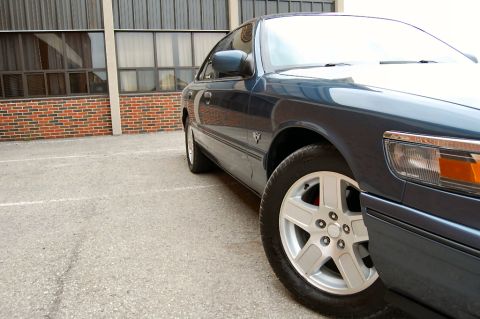 1985 Mercury Grand Marquis - A dark blue sedan is parked in a lot, with a brick building in the background. The vehicle's alloy wheels and side profile are visible.