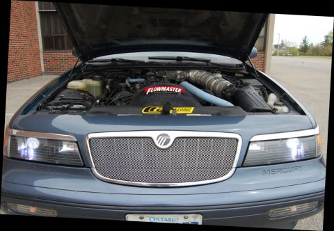 1985 Mercury Grand Marquis - The front of a blue Mercury Grand Marquis is shown with the hood open, revealing the engine and aftermarket intake system.