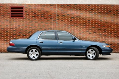 1985 Mercury Grand Marquis - This blue sedan, likely from the late 1990s, is shown from the side in front of a brick building.