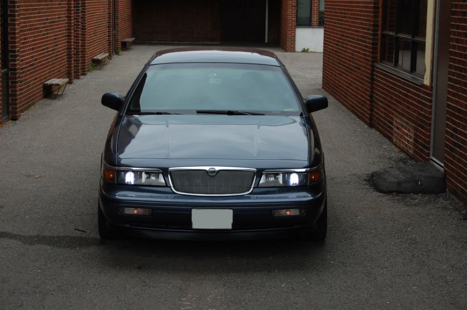 Front view of a dark blue Mercury Grand Marquis with headlights on in a brick alleyway