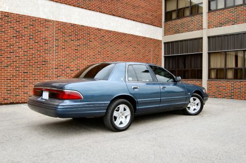 1985 Mercury Grand Marquis - This blue sedan, likely from the late 1990s, is shown from the rear three-quarter angle parked in front of a brick building.