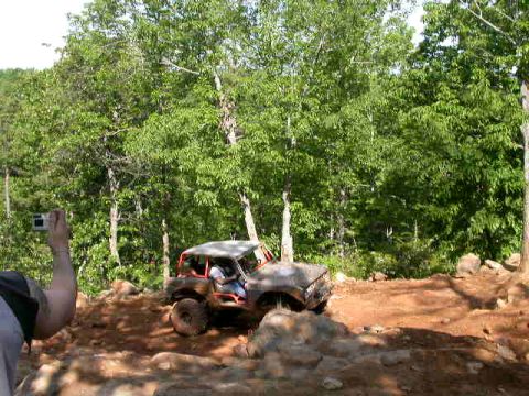 1990 Ford Bronco - An off-road vehicle navigates a challenging dirt terrain with dense trees in the background.