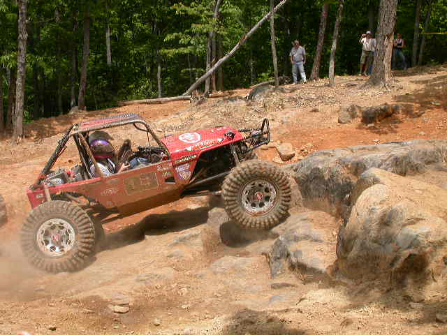 Red off-road buggy climbing over rocks on a dirt trail. Driver wears a helmet