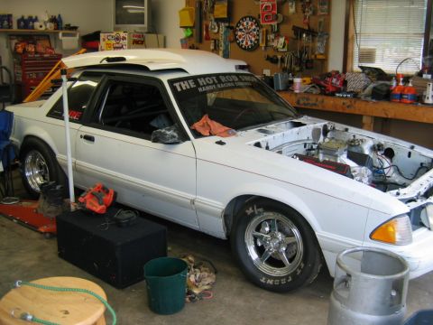 1992 Ford Mustang - A white coupe sits in a garage with its hood open, revealing the engine bay and various tools around it.