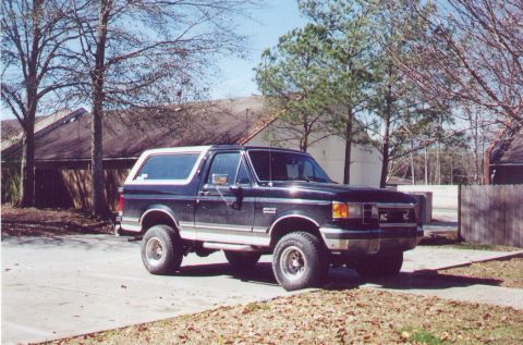 1990 Ford Bronco - A dark blue 1990 Ford Bronco with a white removable top is parked in a lot with trees and buildings in the background.