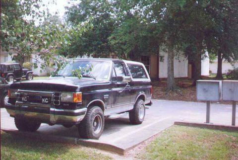 1990 Ford Bronco - A black Ford Bronco is parked in a lot with trees and mailboxes in the background.