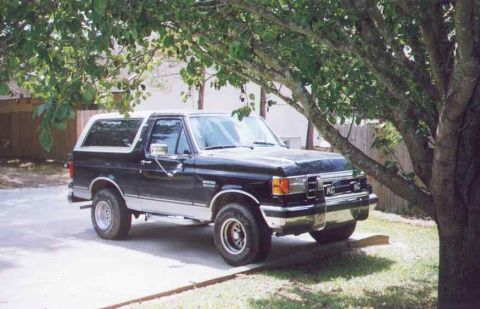 1990 Ford Bronco - A black and silver Ford Bronco is parked on a driveway, partially shaded by tree branches.