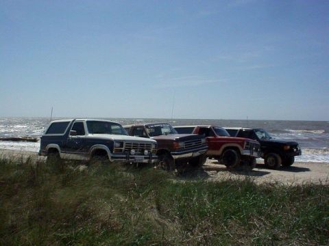 1990 Ford Bronco - A lineup of four SUVs, including a 1990 Ford Bronco, are parked on the sand near the water's edge.