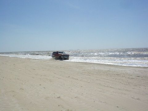 1990 Ford Bronco - A red 1990 Ford Bronco drives along the shoreline with waves crashing around it.
