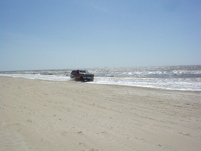 Red 1990 Ford Bronco driving on wet sand near ocean waves