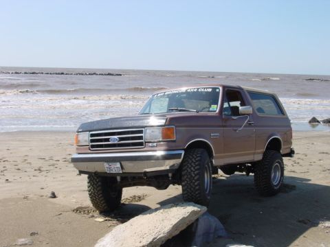 1990 Ford Bronco - A lifted Ford Bronco sits on a beach with the ocean in the background.
