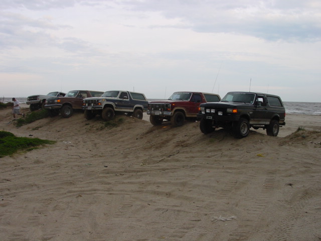 Five Ford Broncos parked on a sandy beach, with the ocean visible.