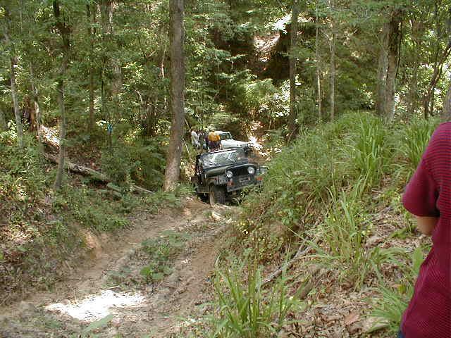 Jeep driving up a muddy, rutted trail in a wooded area.