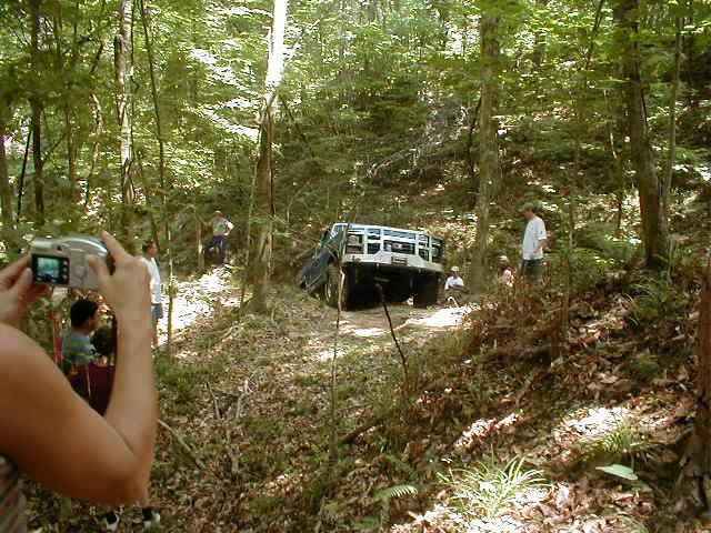 White Ford Bronco tilted on its side in a wooded area with people watching.