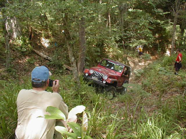 Red Jeep Wrangler driving on a wooded trail with a person taking a photo.