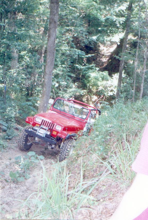 Red Jeep Wrangler driving off-road on a dirt path in a wooded area.